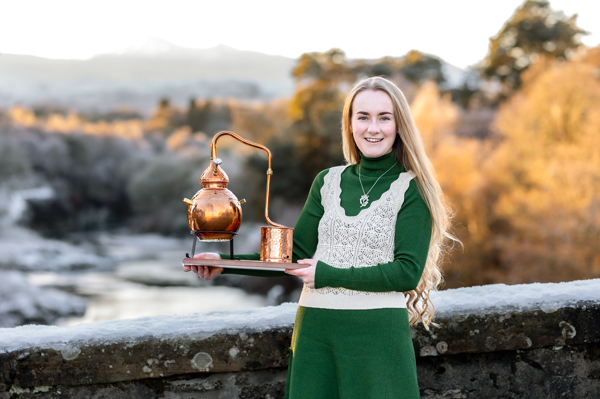 Woman in green dress holding small copper still in front of a wall