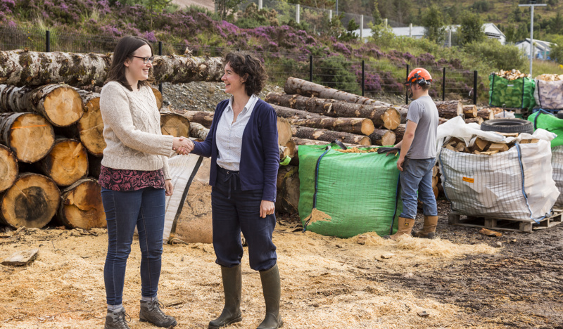 Two women shaking hands in front of a stack of timber